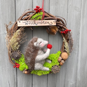 May include: A decorative wreath made of brown twigs, featuring a needle-felted hedgehog holding a red berry. The wreath is adorned with green moss, pinecones, walnuts, and red berries, and includes a wooden sign with the text Willkommen.