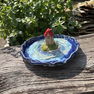 Könnte beinhalten: Eine Keramikschale mit einem skulptierten Miniaturhaus mit rotem Dach, das auf einer kleinen grünen Insel in der Mitte steht. Die Schale hat einen gewellten, geschwungenen Rand und ist in tiefen Blau- und Weißtönen glasiert, die an Wasser erinnern.