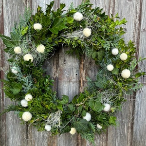 May include: A circular wreath constructed from deep green boxwood and ivy leaves, interspersed with feathery evergreen sprigs. The foliage is accented by numerous small, round, off-white felted wool spheres and thin, wispy white fibers.