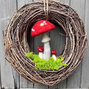 May include: A handcrafted grapevine wreath with a large red mushroom and two smaller mushrooms. The mushrooms have white spots and are set on a bed of green moss. The wreath is suspended by a natural twine.