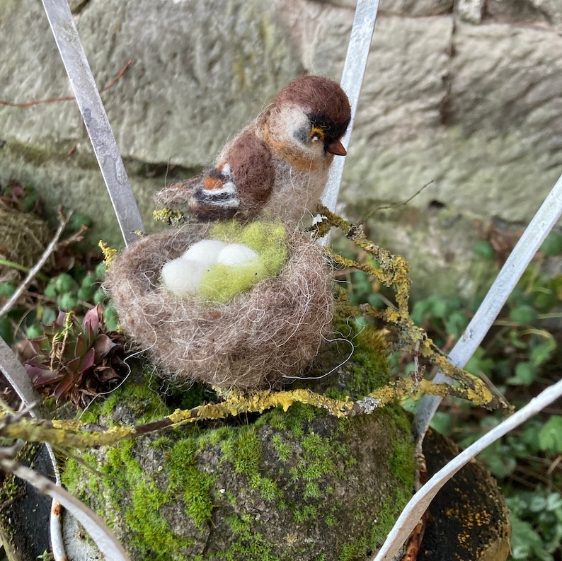 May include: A brown and white felt bird sitting in a brown felt nest with two white eggs. The nest is on a mossy rock with a metal background.