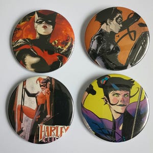 May include: Four round buttons featuring comic book characters. The buttons are black with a silver rim. The characters are Harley Quinn, Batgirl, Catwoman, and a character with a purple suit and a yellow background.