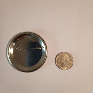 May include: The back of a silver button with a pin on the back. The button is about the size of a US quarter.