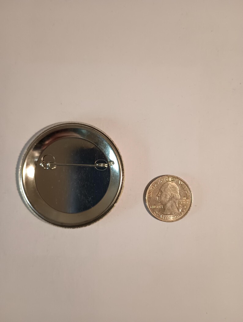 May include: The back of a silver button with a pin on the back. The button is about the size of a US quarter.
