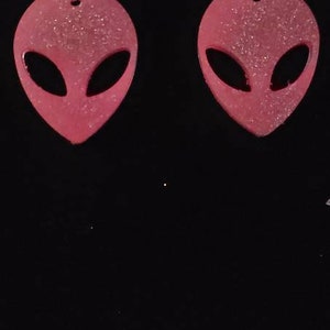 Op de afbeelding: Een paar roze alien-vormige oorbellen met zilveren haakjes. De oorbellen zijn gemaakt van een glinsterend materiaal.