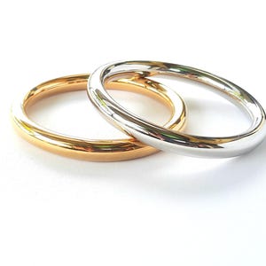May include: Two metal bangles, one gold and one silver, are shown on a white surface. The circular bangles have a smooth, polished finish, reflecting light. The gold bangle is on the left, and the silver bangle is on the right.