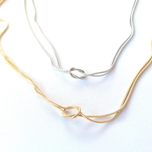 May include: Two delicate necklaces, one gold and one silver, are displayed against a white background. Each necklace features a knot design at the centre. The gold necklace is on the bottom left, and the silver necklace is on the top right.