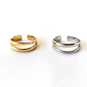 May include: Two adjustable rings, one gold and one silver, are shown on a white surface. Each ring has a double-band design with an open front. The rings are simple and elegant, suitable for everyday wear.