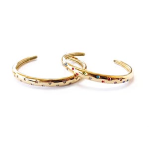 May include: Two gold-toned open cuff bracelets featuring a polished finish and embedded with small, multi-coloured star-shaped accents. The stars are arranged along the outer surface of each circular band, showcasing various colours including red, blue, and clear.