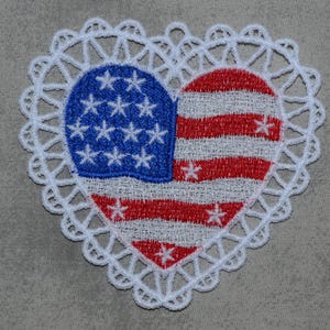 May include: Embroidered heart-shaped patch featuring the American flag design. The blue field with white stars and red and white stripes is surrounded by a white lace trim. Patriotic themed.