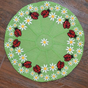 May include: A circular green fabric mat featuring an embroidered pattern of white daisies with yellow centers and red ladybugs with black spots arranged in a ring around the perimeter and a single white daisy centered in the middle of the mat.