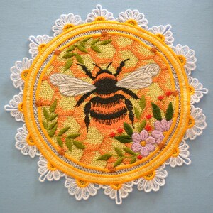 May include: A circular embroidered patch featuring a central bumblebee with black and orange stripes and grey wings. The background is a yellow and orange honeycomb pattern, surrounded by green leaves, small red berries, and purple flowers.