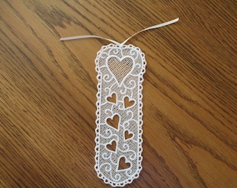 May include: White lace bookmark with a delicate design of hearts and swirls. The bookmark has a white ribbon attached to the top.