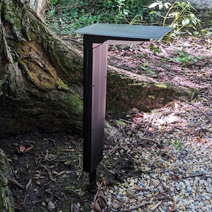 May include: A black metal outdoor light fixture with a rectangular shade mounted on a post. The light is installed in a garden setting with gravel and a tree trunk in the background.