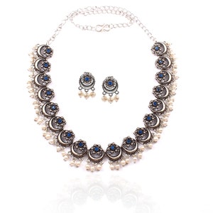 May include: A silver-toned necklace and matching earrings set. The necklace features crescent-shaped pendants with blue gemstone centers and dangling white pearls. The earrings mirror the design. The jewelry is displayed on a white background.
