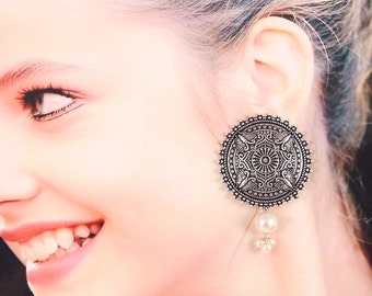 May include: Silver tone earrings with a round, ornate design and a pearl drop. The earrings are silver with a detailed, intricate pattern.