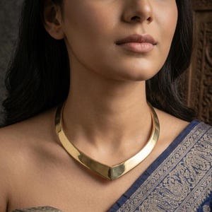 May include: A gold-coloured collar necklace with a V-shaped design. The necklace is worn around the neck and sits on the collarbone. The woman is wearing a blue patterned garment.