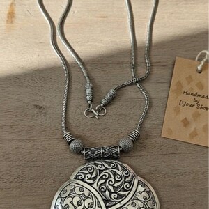 May include: Silver-toned necklace with a large, ornate pendant. The pendant features an intricate floral design. The necklace has a chain-like structure with a clasp closure. A tag with text is visible in the background. The necklace is on a wooden surface.
