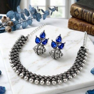 May include: A silver-toned necklace and matching earrings set. The necklace features a beaded design with a series of small, round beads. The earrings have a teardrop-shaped blue gemstone design. The jewelry is displayed on a white surface.