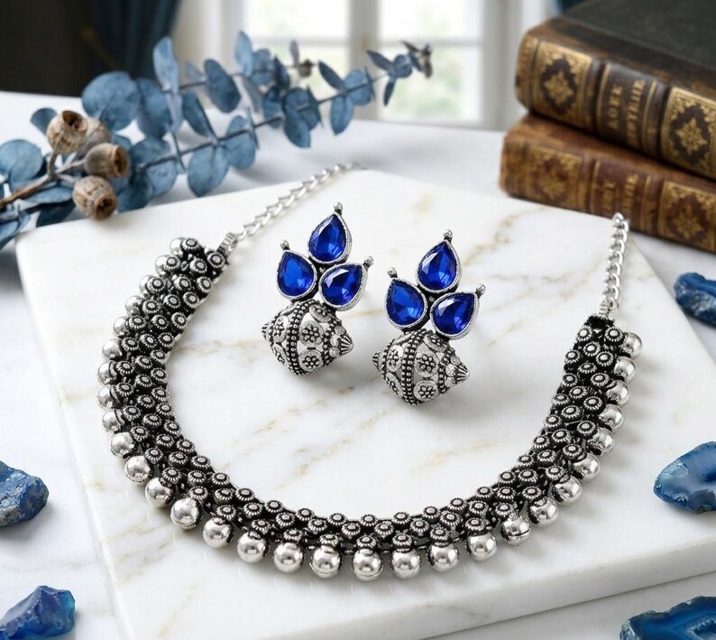 May include: A silver-toned necklace and matching earrings set. The necklace features a beaded design with a series of small, round beads. The earrings have a teardrop-shaped blue gemstone design. The jewelry is displayed on a white surface.