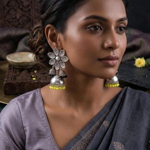 May include: Silver-toned floral earrings with a layered design. The earrings feature a flower-shaped top, two bell-shaped dangles, and yellow bead accents. The earrings are worn by a person wearing a gray top.
