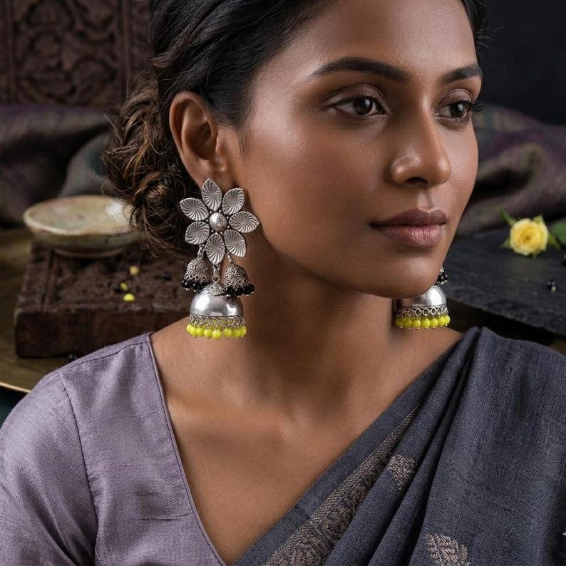 May include: Silver-toned floral earrings with a layered design. The earrings feature a flower-shaped top, two bell-shaped dangles, and yellow bead accents. The earrings are worn by a person wearing a gray top.
