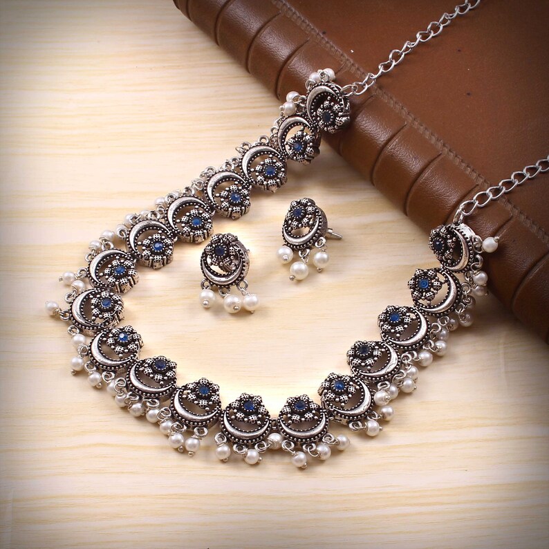 May include: A silver-toned necklace and earring set. The necklace and earrings feature a crescent moon design with blue center stones and small white pearl accents. The jewelry is displayed on a light wood surface.