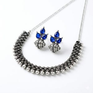 May include: A silver-toned necklace and matching earrings set. The necklace features a beaded design with a central section of small, round beads. The earrings have a cluster of three blue teardrop-shaped stones. The jewelry is set against a white background.