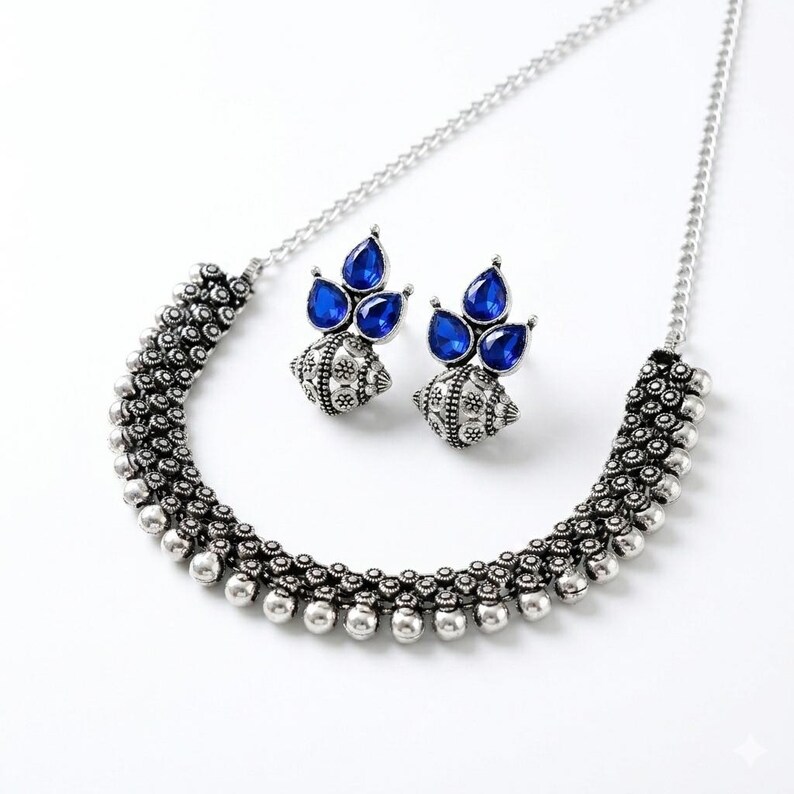 May include: A silver-toned necklace and matching earrings set. The necklace features a beaded design with a central section of small, round beads. The earrings have a cluster of three blue teardrop-shaped stones. The jewelry is set against a white background.