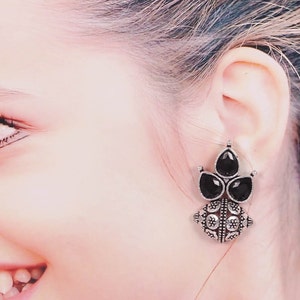 May include: Silver tone earrings with black gemstone accents. The earrings feature a round, ornate design with a cluster of three black gemstones.