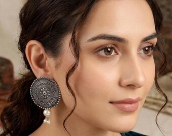 May include: A large, circular, oxidized silver-tone earring featuring intricate, concentric etched patterns and a central floral motif. A single, smooth, cream-colored pearl bead hangs from the bottom, accented by two smaller, delicate pearl beads.