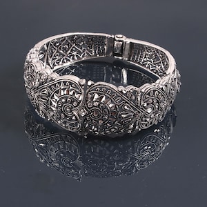 Peut inclure: Bracelet articulé argenté orné de motifs floraux et géométriques complexes. Le bracelet présente un design détaillé avec un fermoir sécurisé. Le bijou est présenté sur une surface réfléchissante, accentuant son aspect orné.