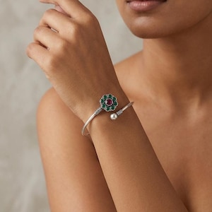 May include: A silver-toned open cuff bracelet featuring a floral-shaped centerpiece with a central round red stone surrounded by small green stones. The band has a textured ribbed detail near the flower and a smooth silver ball at the other end.