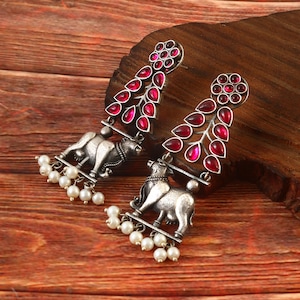 May include: A pair of silver-toned earrings. Each earring features a detailed bull figure, a cascade of white pearls, and a teardrop-shaped red gemstone design. The earrings are displayed on a wooden surface.