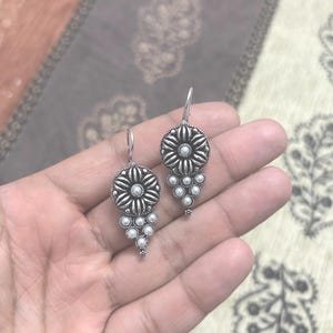 以下が含まれることがあります： 花柄のデザインと白い真珠のアクセントが付いたシルバーのドロップイヤリング