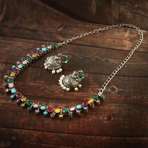 May include: A silver-toned necklace and matching earrings set. The necklace and earrings feature colorful gemstones in various colors, including red, green, yellow, and blue. The earrings are a dome shape with small white beads.