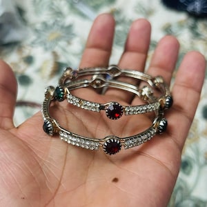 May include: Two silver-toned metal bangles featuring rows of small clear rhinestones and larger circular inset stones in deep red, green, and black colors. The bangles have a polished metallic finish and a circular shape with alternating stone settings.