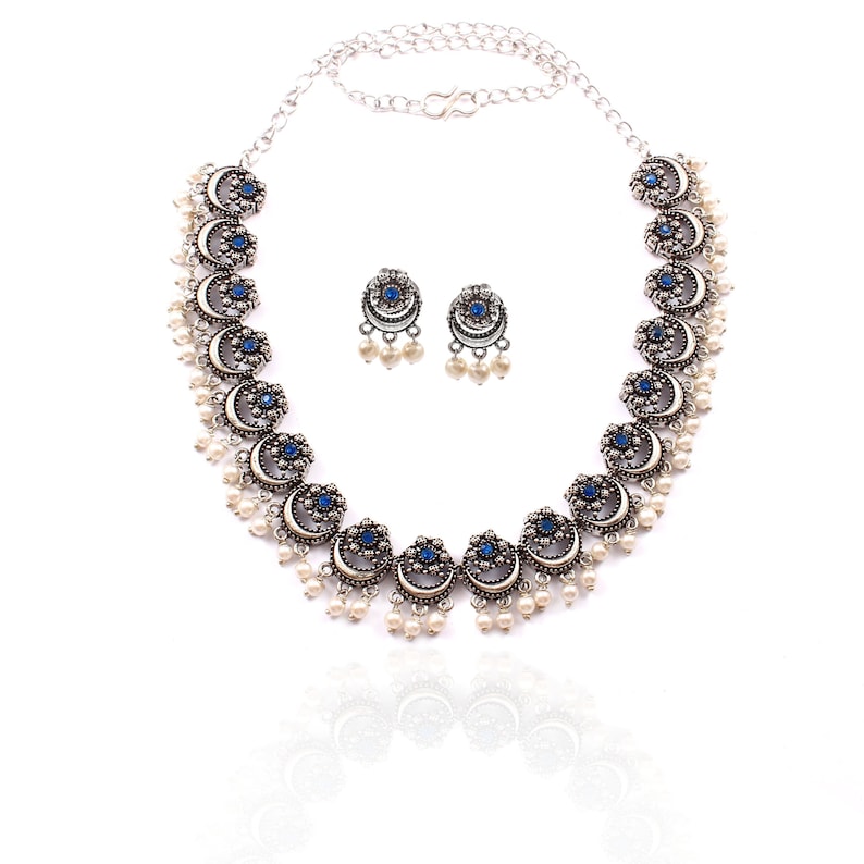 May include: A silver-toned necklace and matching earrings set. The necklace features crescent-shaped pendants with blue gemstone centers and dangling white pearls. The earrings mirror the design. The jewelry is displayed on a white background.