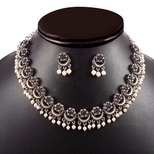 May include: A silver-toned necklace and earring set. The necklace features crescent-shaped pendants with blue center stones and dangling white pearls. The matching earrings have a similar design. The jewelry is displayed on a black mannequin.