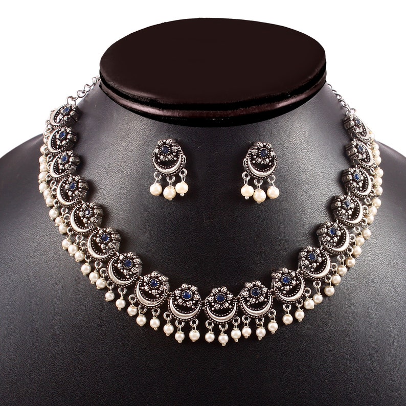 May include: A silver-toned necklace and earring set. The necklace features crescent-shaped pendants with blue center stones and dangling white pearls. The matching earrings have a similar design. The jewelry is displayed on a black mannequin.