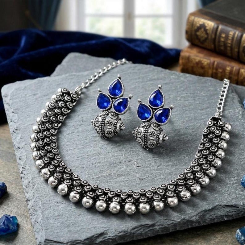 May include: A silver-toned necklace and matching earrings set. The necklace and earrings feature a detailed, ornate design with small, round accents. The earrings have three teardrop-shaped, blue gemstones. The jewelry set is displayed on a gray surface.