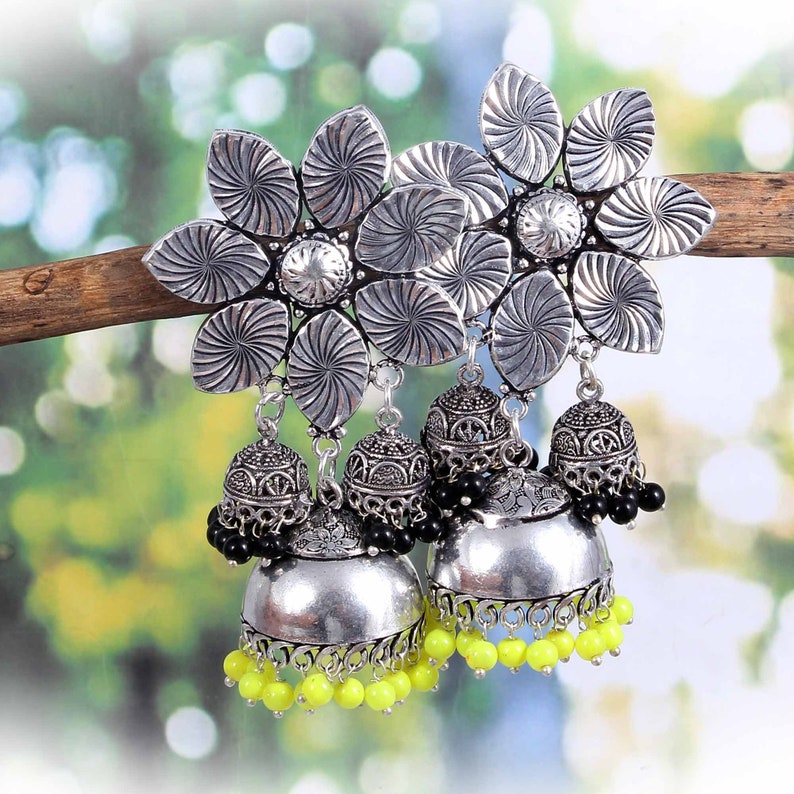 May include: Silver-toned earrings with a floral design at the top. The earrings have a bell-shaped dangle with black and neon yellow bead accents. The earrings are displayed on a wooden branch against a blurred background.