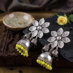 May include: Silver-toned flower earrings with a detailed design. Each earring features a large flower with radiating lines, a small bell, and a cluster of yellow beads. The earrings are displayed on a dark surface.