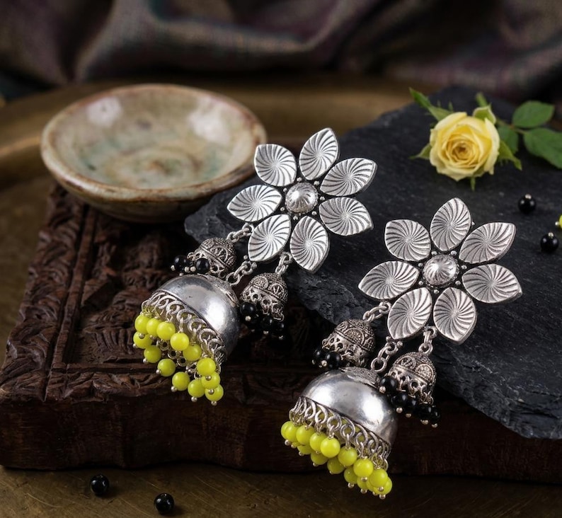 May include: Silver-toned flower earrings with a detailed design. Each earring features a large flower with radiating lines, a small bell, and a cluster of yellow beads. The earrings are displayed on a dark surface.