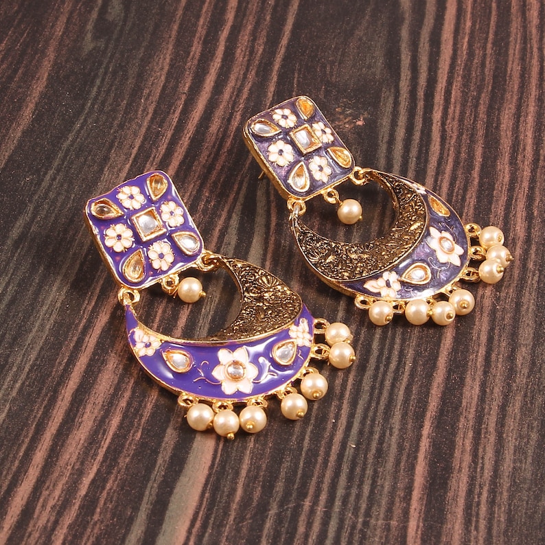 May include: A pair of purple and gold earrings with white floral designs and pearl accents. The earrings are shaped like a crescent moon with a square pendant at the top.