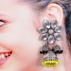 May include: Silver-toned flower earrings with a central clear gem and dangling bell-shaped charms. The charms feature black and yellow beads, creating a striking contrast. The earrings are designed with intricate details.