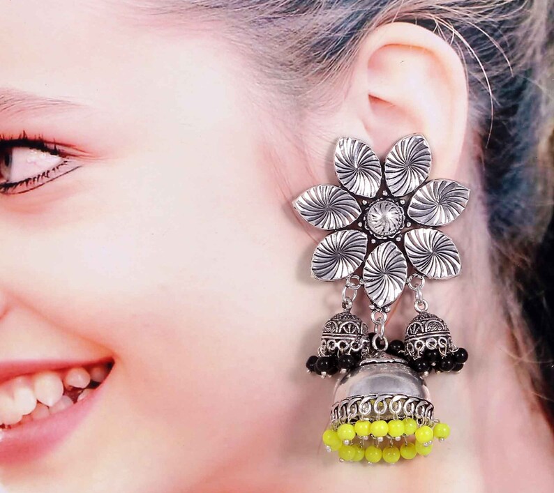 May include: Silver-toned flower earrings with a central clear gem and dangling bell-shaped charms. The charms feature black and yellow beads, creating a striking contrast. The earrings are designed with intricate details.