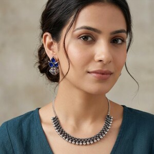 May include: A silver-toned necklace and earring set. The necklace features a curved design with small, round beads and a chain. The earrings have a floral design with blue teardrop-shaped stones. The jewelry is worn against a teal-colored top.