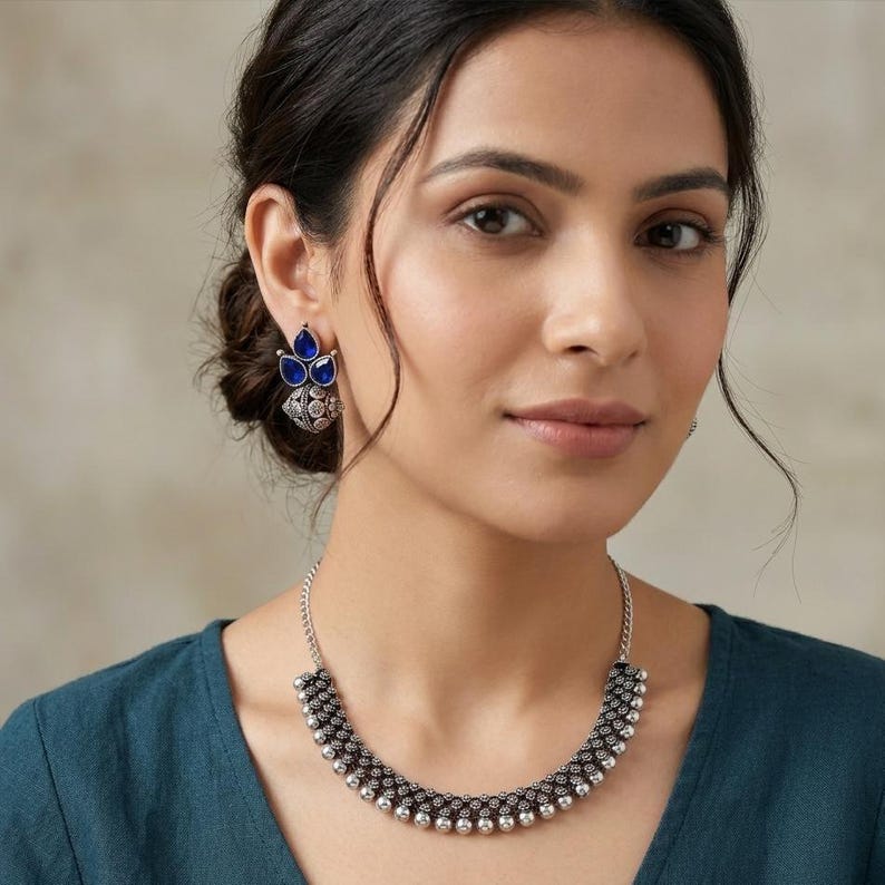 May include: A silver-toned necklace and earring set. The necklace features a curved design with small, round beads and a chain. The earrings have a floral design with blue teardrop-shaped stones. The jewelry is worn against a teal-colored top.