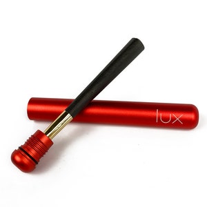 Puede incluir: Un tubo de metal rojo con un palo de metal negro y dorado en su interior. El tubo tiene la palabra "lux" impresa en él.